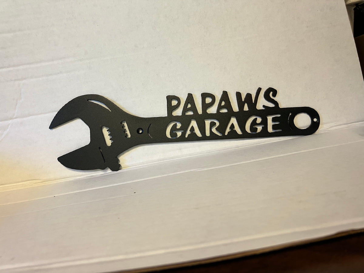 A black metal wrench-shaped sign with the words 'PAPAWS GARAGE' cut into the handle, resting on a white cardboard box.