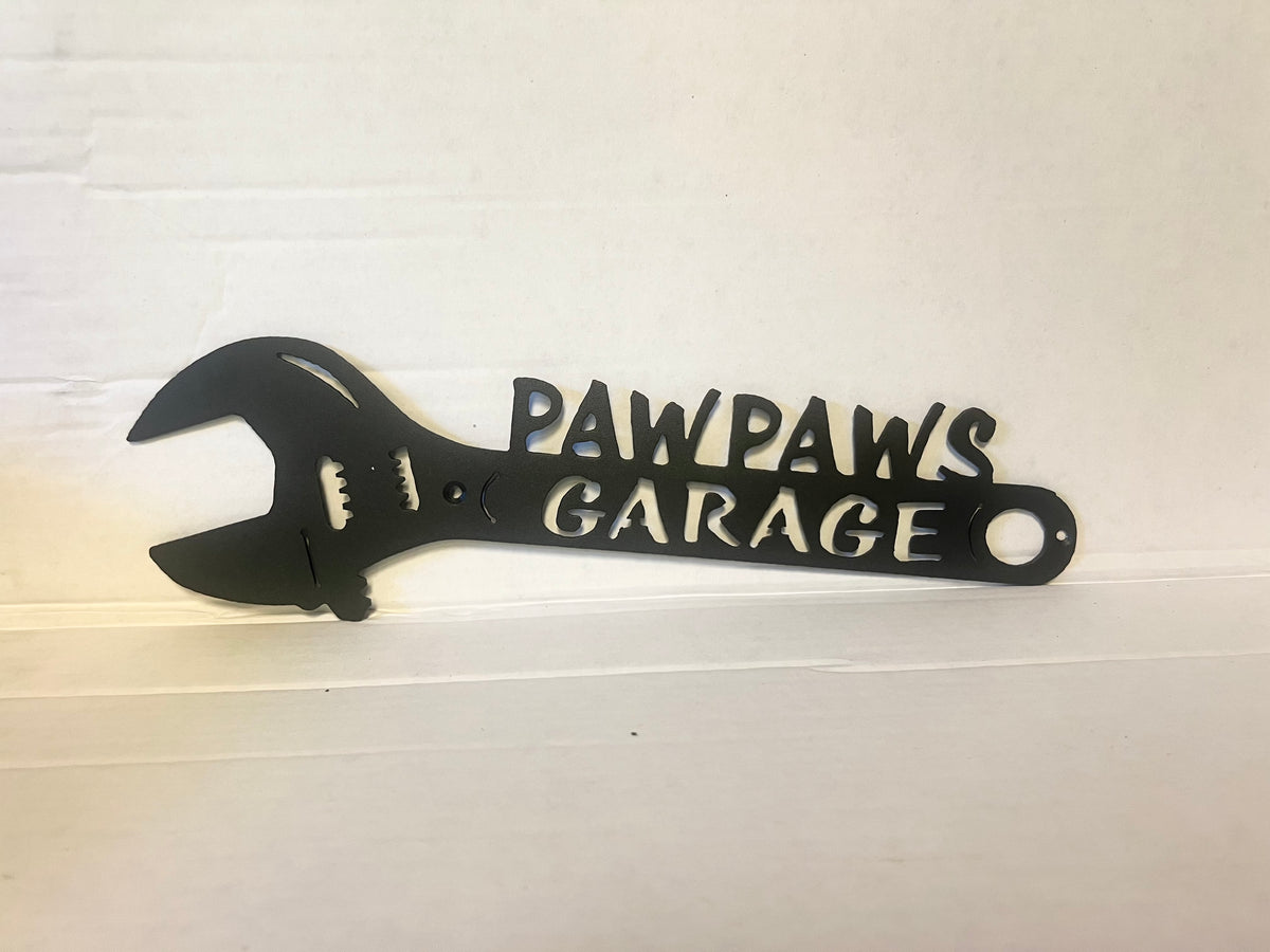 A black metal wrench-shaped wall sign with the words 'PAWPAWS GARAGE' cut out, displayed against a white painted wooden background.