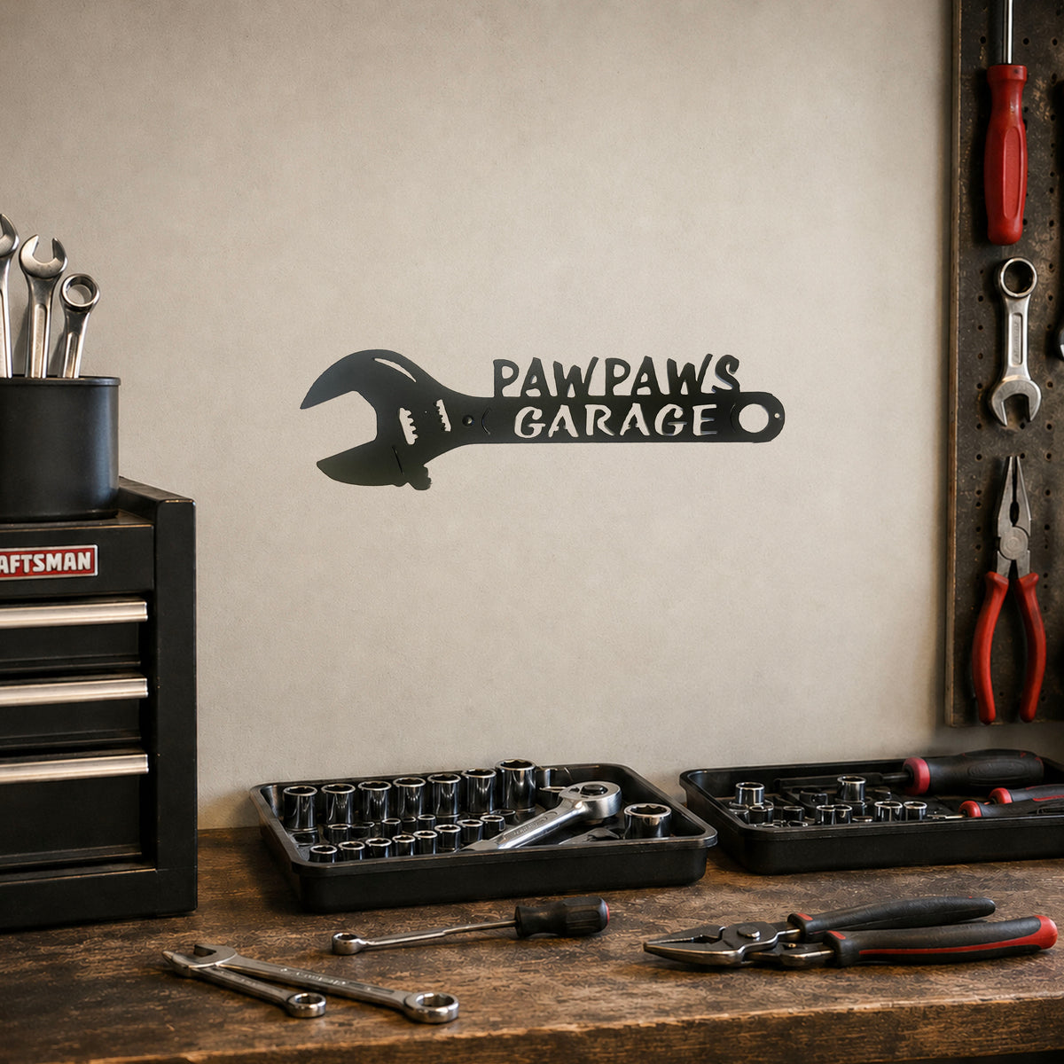 A black metal wall decal shaped like a wrench with the text 'PAWPAWS GARAGE' mounted on a beige wall in a garage setting, with tool trays, wrenches, pliers, and a Craftsman tool chest on a wooden workbench below.
