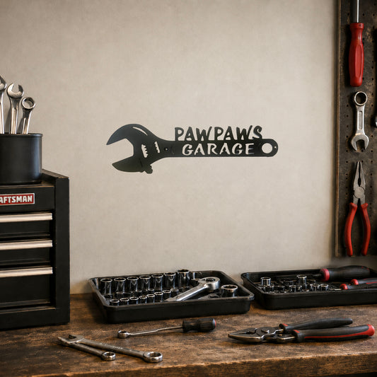 A black metal wall decal shaped like a wrench with the text 'PAWPAWS GARAGE' mounted on a beige wall in a garage setting, with tool trays, wrenches, pliers, and a Craftsman tool chest on a wooden workbench below.