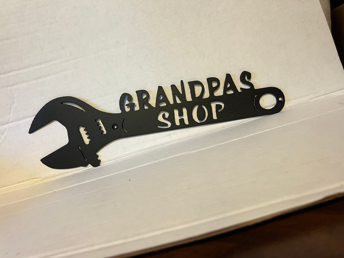 A black metal sign shaped like an adjustable wrench with the words 'GRANDPA'S SHOP' cut out, placed on a white surface.