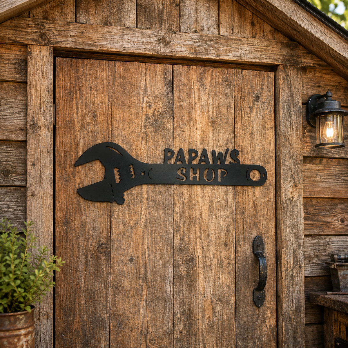 A rustic wooden door with a black metal wrench-shaped sign reading 'PAPAWS SHOP' mounted on the door, set in a weathered wood shed with a wall-mounted lantern light.