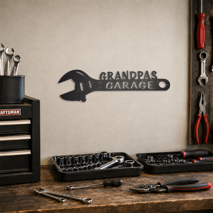 A black metal wrench-shaped wall sign with the words 'GRANDPA GARAGE' mounted on a beige wall in a garage workshop setting, with tools and a Craftsman toolbox on a wooden workbench below.