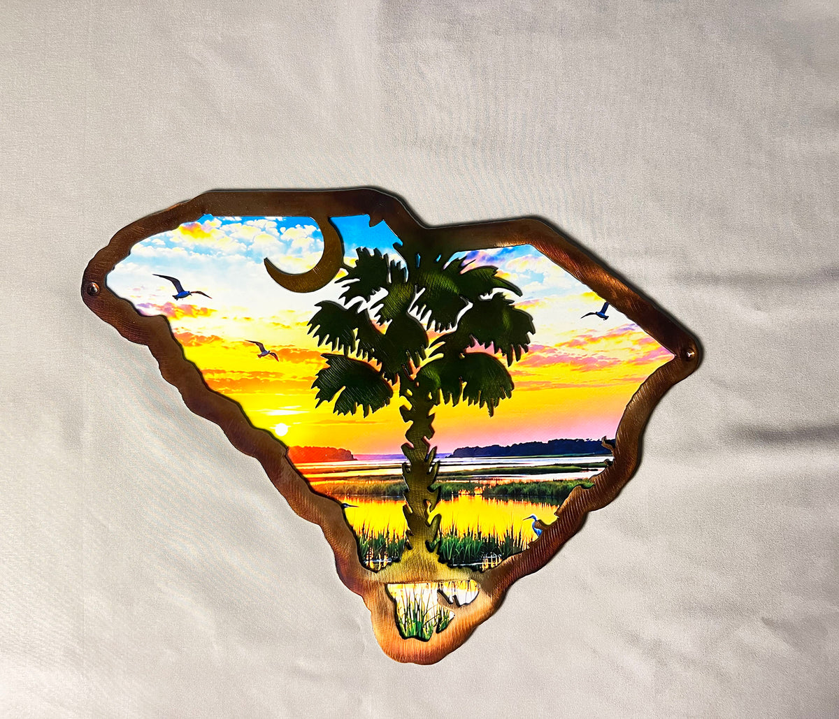 A metal wall sign shaped like the state of South Carolina, featuring a hand-painted sunset landscape with a palm tree, birds, and a crescent moon, displayed on a white fabric background.