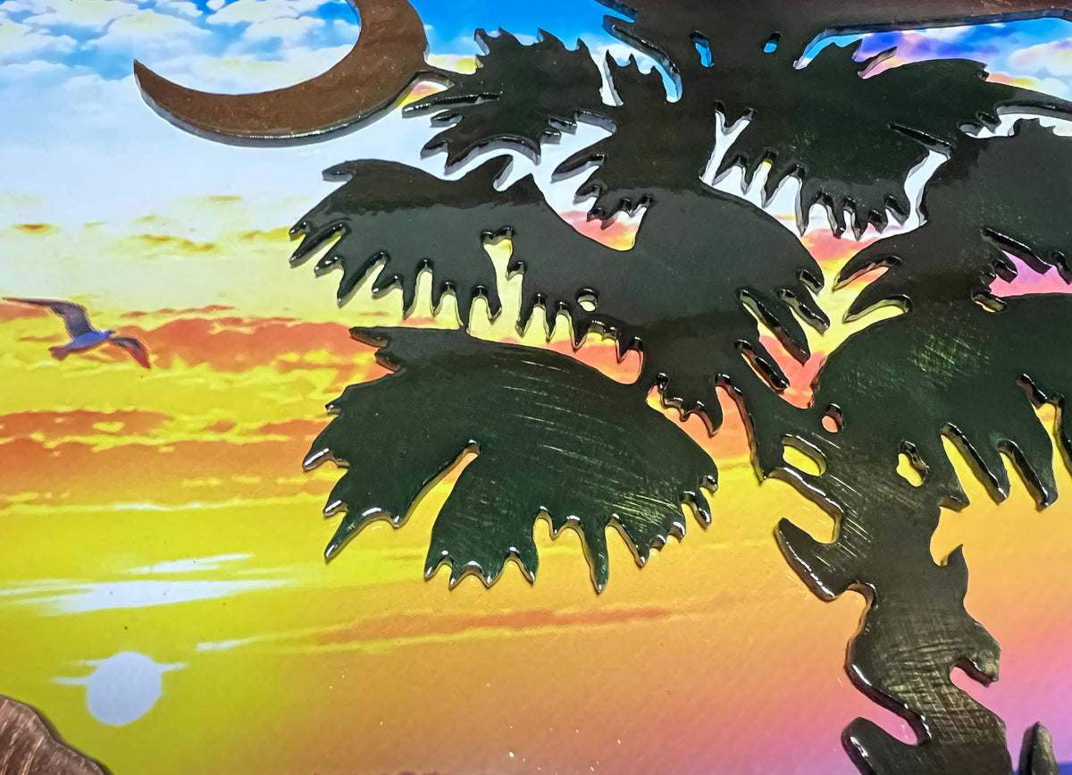 Close-up of a decorative metal wall art piece featuring a dark silhouette of a palm tree in front of a colorful sunset sky with a crescent moon and bird.
