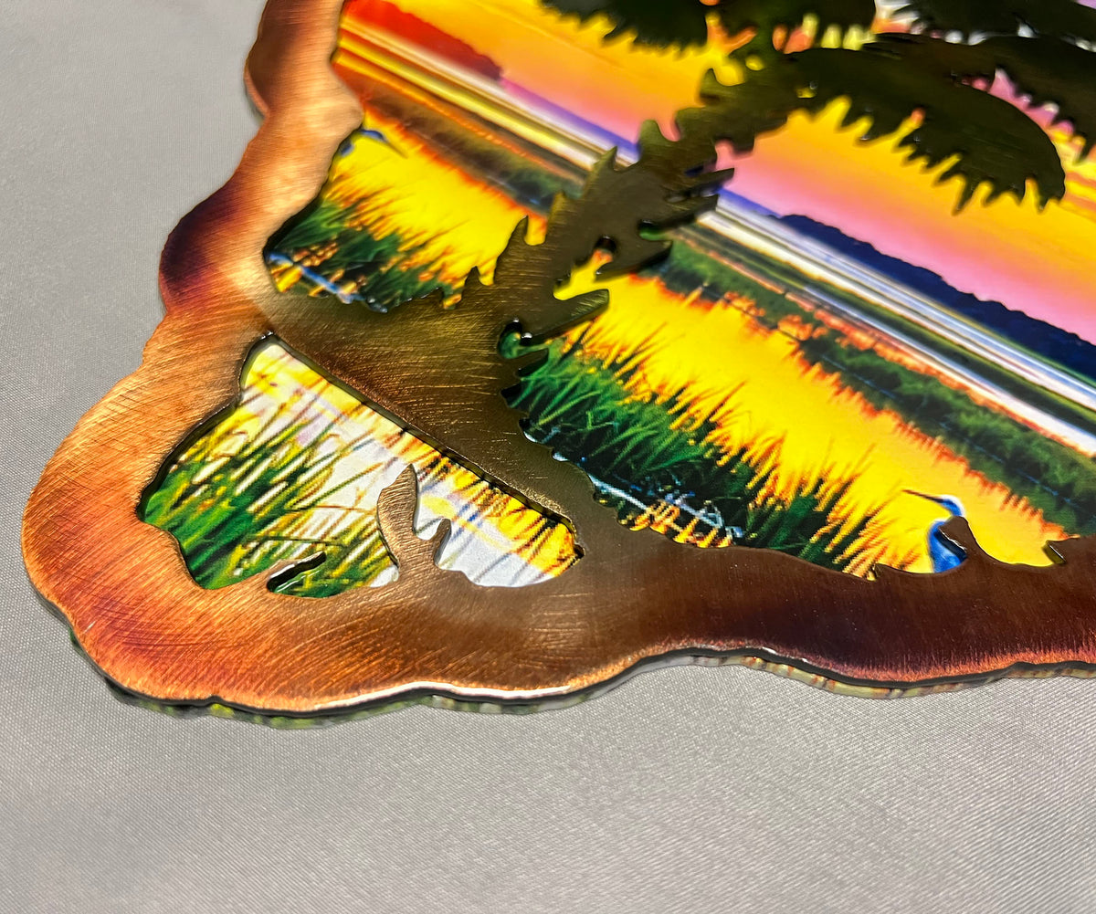 A close-up of a laser-cut metal wall art piece featuring a colorful sunset landscape with a bird, shown on a neutral gray surface.