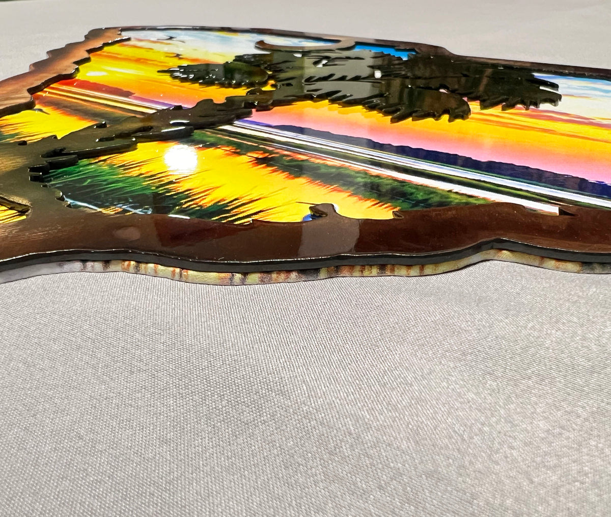 A close-up of a decorative metal art piece featuring a colorful sunset landscape design with layered cutouts, displayed on a light gray textured fabric background.