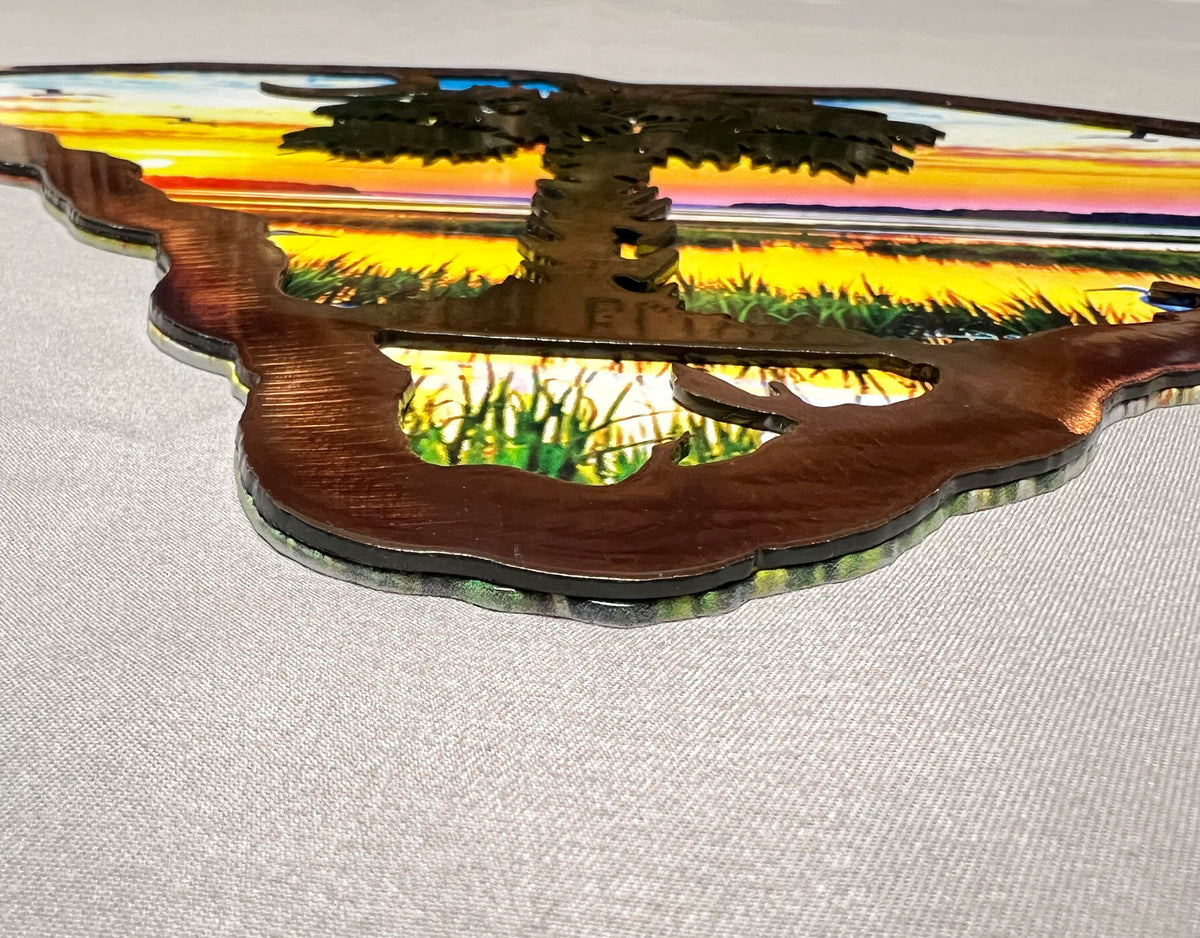 A close-up of a laser-cut wooden wall art piece featuring a tree and grassy landscape with a sunset sky, displayed on a light gray fabric background.