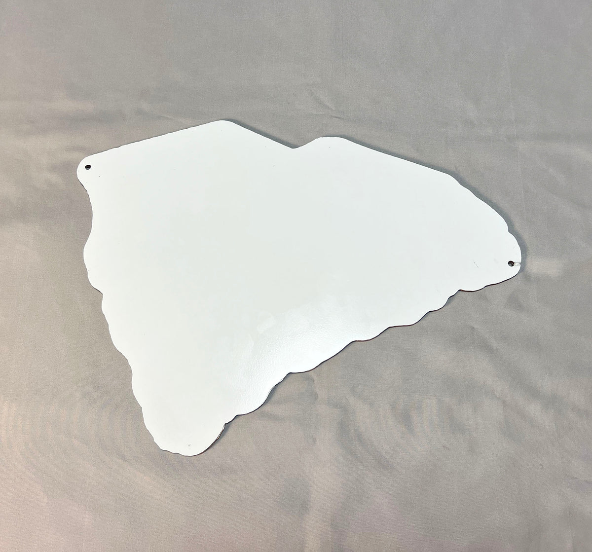 A white, irregularly shaped metal cutout with two pre-drilled holes, displayed on a textured gray fabric background.