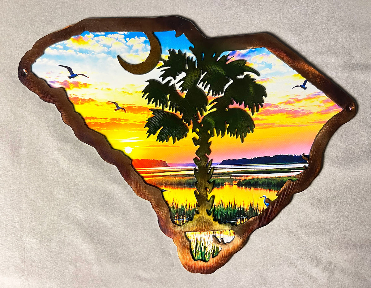 A decorative metal wall sign shaped like the state of South Carolina, featuring a colorful sunset landscape with a palm tree, birds, and a crescent moon, mounted on a light fabric background.