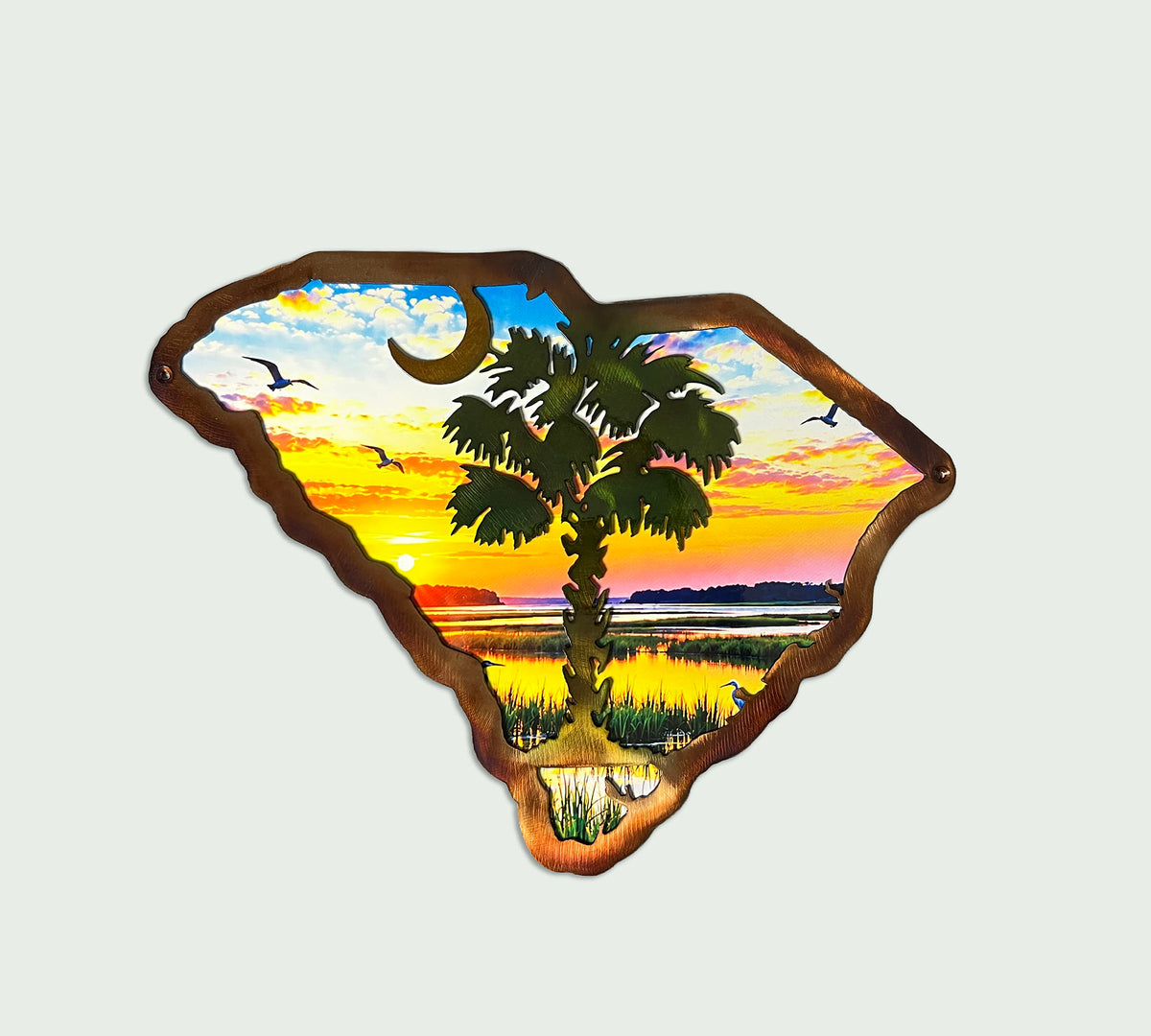 A metal wall sign shaped like the South Carolina state, featuring a painted sunset landscape with a palm tree, birds, and a crescent moon, mounted on a light gray background.
