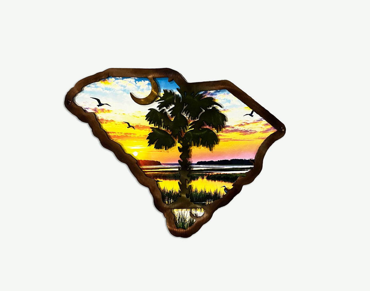 A decorative wall art piece shaped like the state of South Carolina, featuring a sunset landscape with a palm tree, birds, water, and a crescent moon, outlined in dark brown wood against a light gray background.
