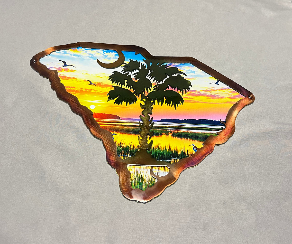 A metal wall art piece shaped like the state of South Carolina, featuring a sunset marsh landscape with a palm tree, birds, and a crescent moon, displayed on a light gray fabric background.