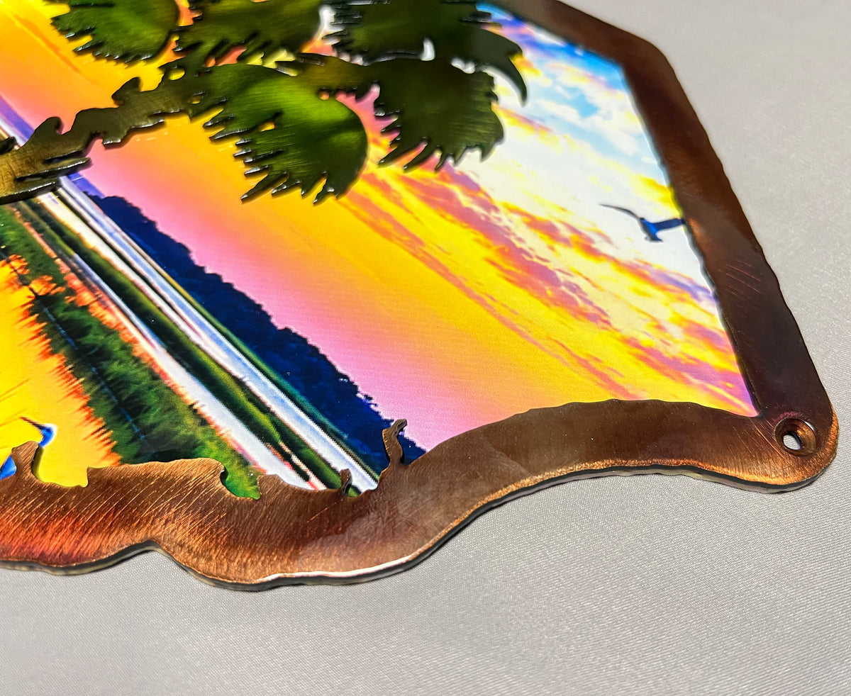 A close-up of a decorative metal wall sign featuring a colorful sunset landscape with palm tree branches layered over the scene. The sign has a wavy copper-toned metal border and a small hanging hole in the upper right corner, photographed on a light gray surface.