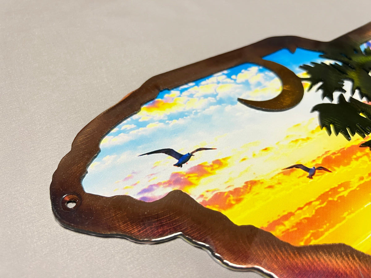 A close-up of a decorative metal wall sign with an irregular cutout border, featuring a sunset sky scene with birds, clouds, a crescent moon, and palm leaves. The sign is photographed on a light neutral surface.