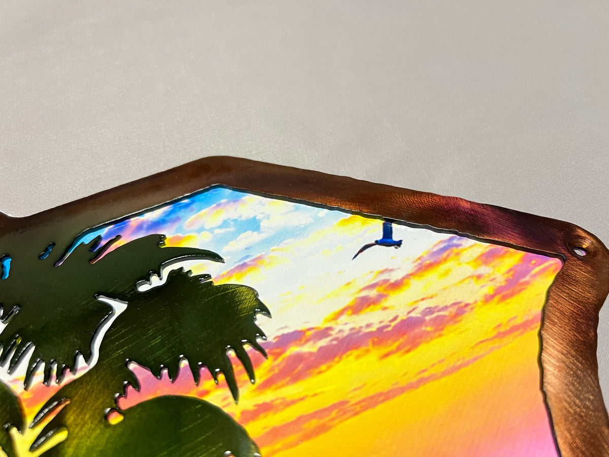 A close-up of a metal wall art piece featuring a colorful sunset sky with palm tree silhouettes and a bird, set against a plain light gray background.